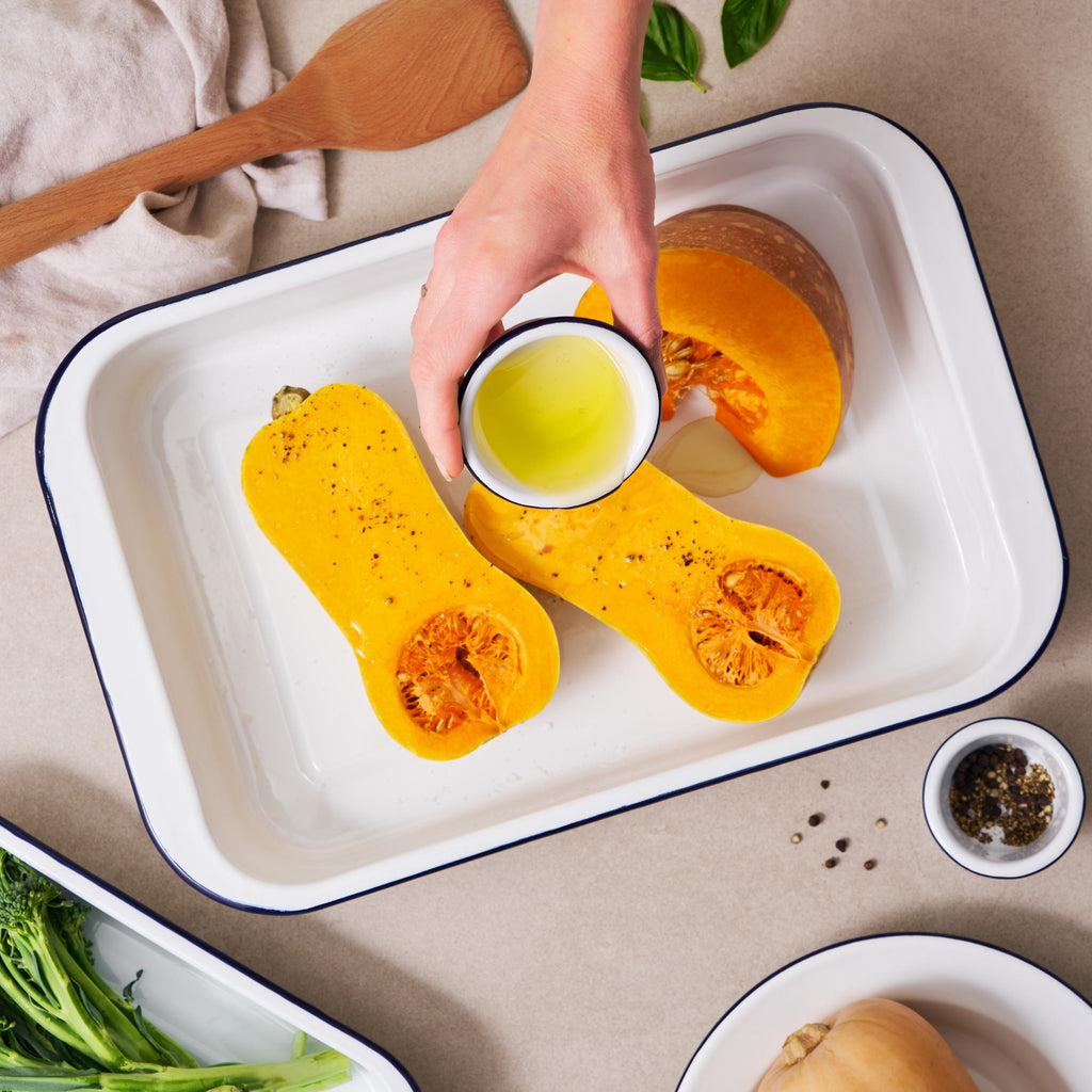 A top-down view of a hand holding a small cup of olive oil over a white enamel baking tray containing two sliced butternut squash halves, with other cooking ingredients and vegetables arranged around it on a beige surface.